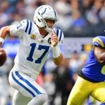 Daniel Jones in einem weißen Trikot der Indianapolis Colts, Nummer 17, läuft mit dem Ball, während er von einem Spieler der Los Angeles Rams während eines NFL-Spiels der regulären Saison 2025 verfolgt wird. Der Hintergrund zeigt eine unscharfe Zuschauermenge und das Spielfeld, wobei zwei der besten Spieler in Aktion zu sehen sind. Diese Beschreibung wurde mit der FootballR KI automatisch generiert.