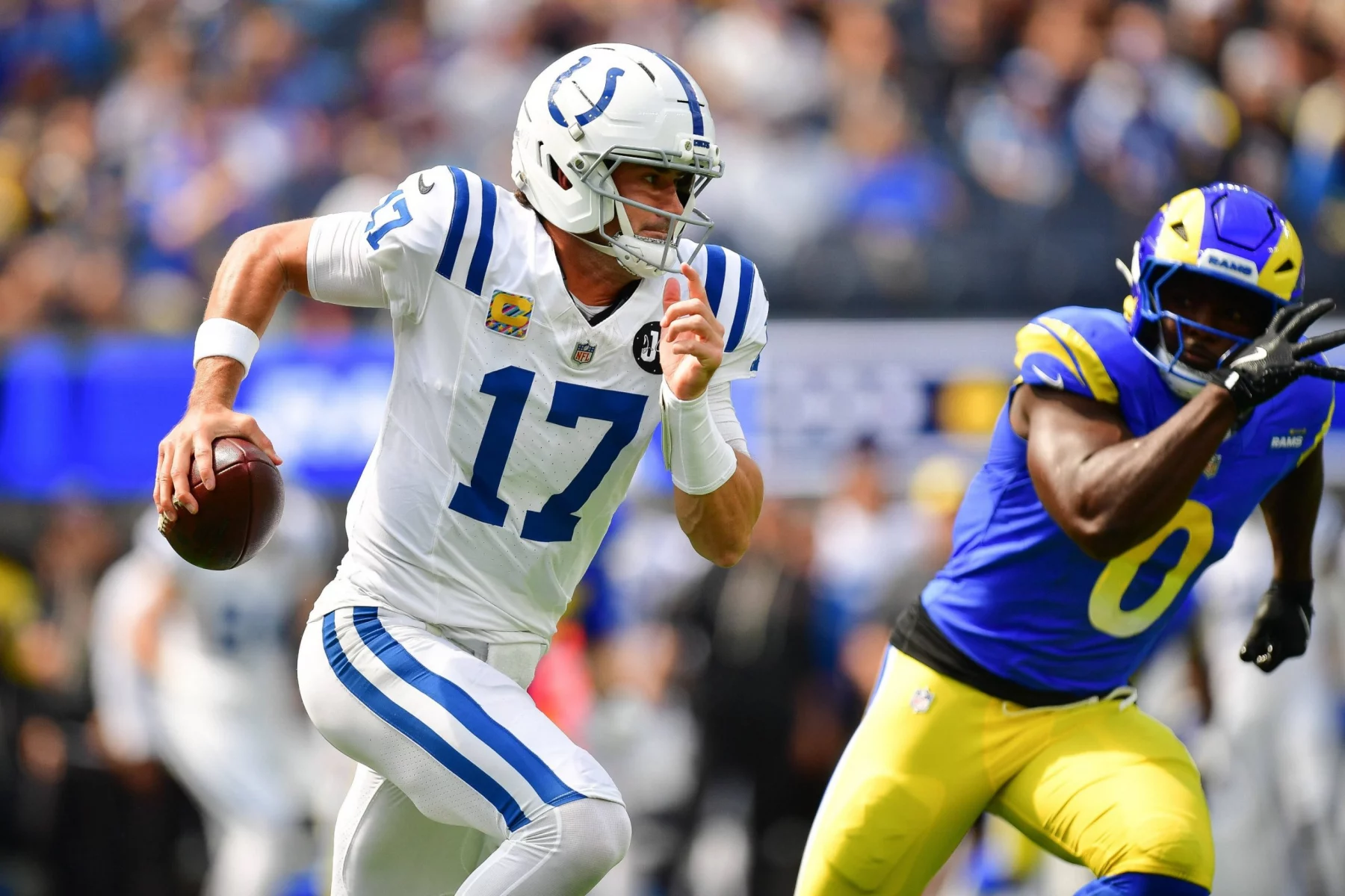 Free Agency 2026 - Daniel Jones in einem weißen Trikot der Indianapolis Colts, Nummer 17, läuft mit dem Ball, während er von einem Spieler der Los Angeles Rams während eines NFL-Spiels der regulären Saison 2025 verfolgt wird. Der Hintergrund zeigt eine unscharfe Zuschauermenge und das Spielfeld, wobei zwei der besten Spieler in Aktion zu sehen sind. Diese Beschreibung wurde mit der FootballR KI automatisch generiert.