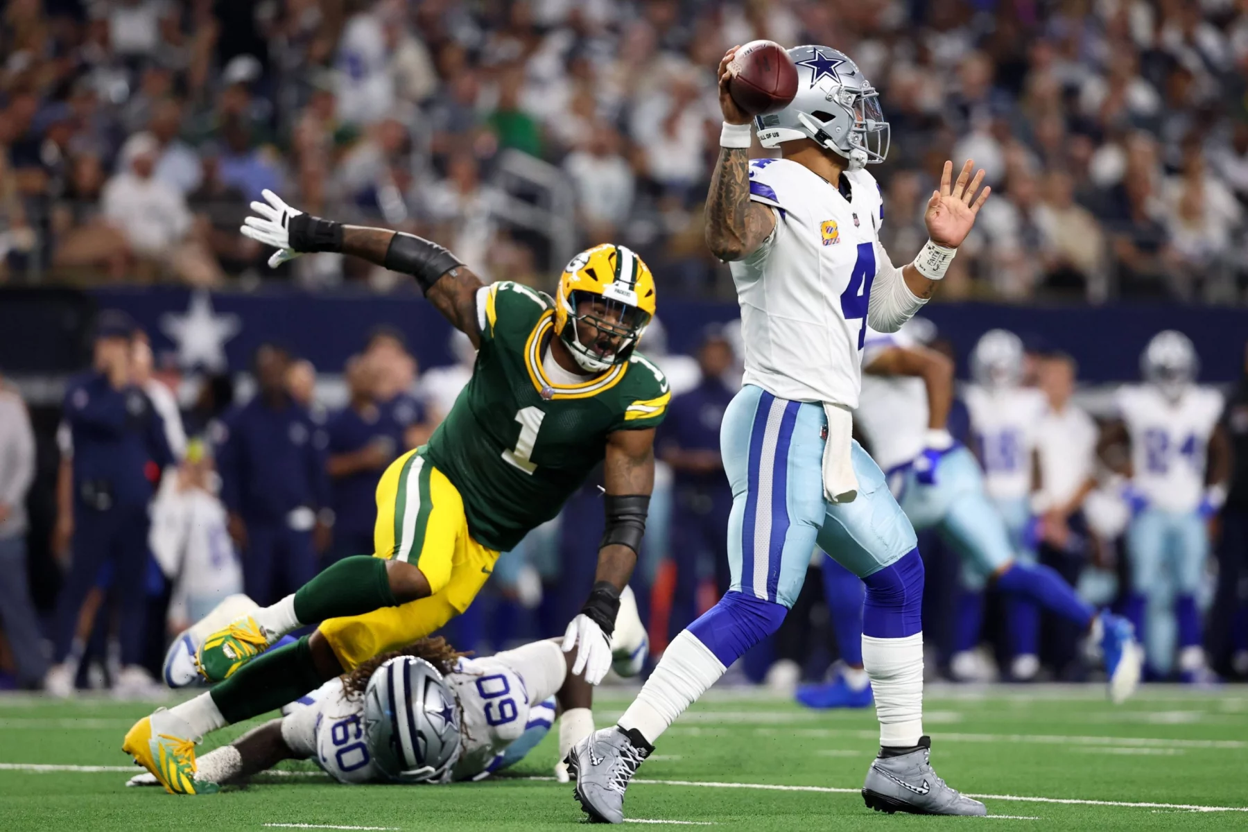 Ein Quarterback der Dallas Cowboys in Weiß und Blau bereitet sich auf den Wurf vor, während ein Verteidiger der Packers in Grün und Gelb auf ihn zustürzt. Andere Spieler und die Zuschauer füllen das Stadion bei diesem spannenden Spiel zwischen Dallas und den Packers. Diese Beschreibung wurde mit der FootballR KI automatisch generiert.
