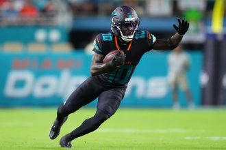 Tyreek Hill, die Nummer 10 der Miami Dolphins, sprintet in schwarzer Uniform, mit Helm und Handschuhen über das grüne Feld. Er beugt sich mit ausgestrecktem Arm nach vorne, während im Hintergrund Fans und "Dolphins"-Schriftzüge verschwimmen - keine Spur von Beinverletzung. Diese Beschreibung wurde mit der FootballR KI automatisch generiert.