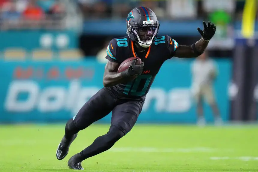 Tyreek Hill, die Nummer 10 der Miami Dolphins, sprintet in schwarzer Uniform, mit Helm und Handschuhen über das grüne Feld. Er beugt sich mit ausgestrecktem Arm nach vorne, während im Hintergrund Fans und "Dolphins"-Schriftzüge verschwimmen - keine Spur von Beinverletzung. Diese Beschreibung wurde mit der FootballR KI automatisch generiert.