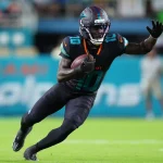 Tyreek Hill, die Nummer 10 der Miami Dolphins, sprintet in schwarzer Uniform, mit Helm und Handschuhen über das grüne Feld. Er beugt sich mit ausgestrecktem Arm nach vorne, während im Hintergrund Fans und "Dolphins"-Schriftzüge verschwimmen - keine Spur von Beinverletzung. Diese Beschreibung wurde mit der FootballR KI automatisch generiert.