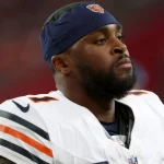 Jaylon Johnson, der eine Chicago Bears-Uniform mit marineblauen und orangefarbenen Streifen und einem passenden Stirnband trägt, steht konzentriert auf dem Spielfeld. Er hat einen kurzen Bart und blickt in die Ferne, wobei der unscharfe Stadionhintergrund seine Präsenz hervorhebt. Diese Beschreibung wurde mit der FootballR KI automatisch generiert.