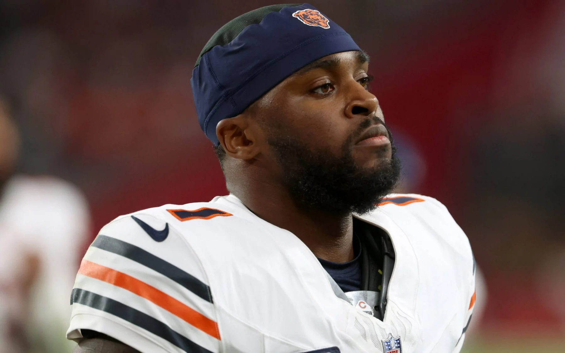 Jaylon Johnson, der eine Chicago Bears-Uniform mit marineblauen und orangefarbenen Streifen und einem passenden Stirnband trägt, steht konzentriert auf dem Spielfeld. Er hat einen kurzen Bart und blickt in die Ferne, wobei der unscharfe Stadionhintergrund seine Präsenz hervorhebt. Diese Beschreibung wurde mit der FootballR KI automatisch generiert.