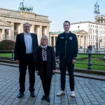 Drei Personen stehen nebeneinander auf einer gepflasterten Fläche in Berlin, mit dem berühmten Brandenburger Tor im Rücken. Sie lächeln und sind sowohl formell als auch leger gekleidet. Es scheint, als würden sie eine der lebhaften Veranstaltungen der Stadt unter einem klaren Himmel genießen. Diese Beschreibung wurde mit der FootballR KI automatisch generiert.