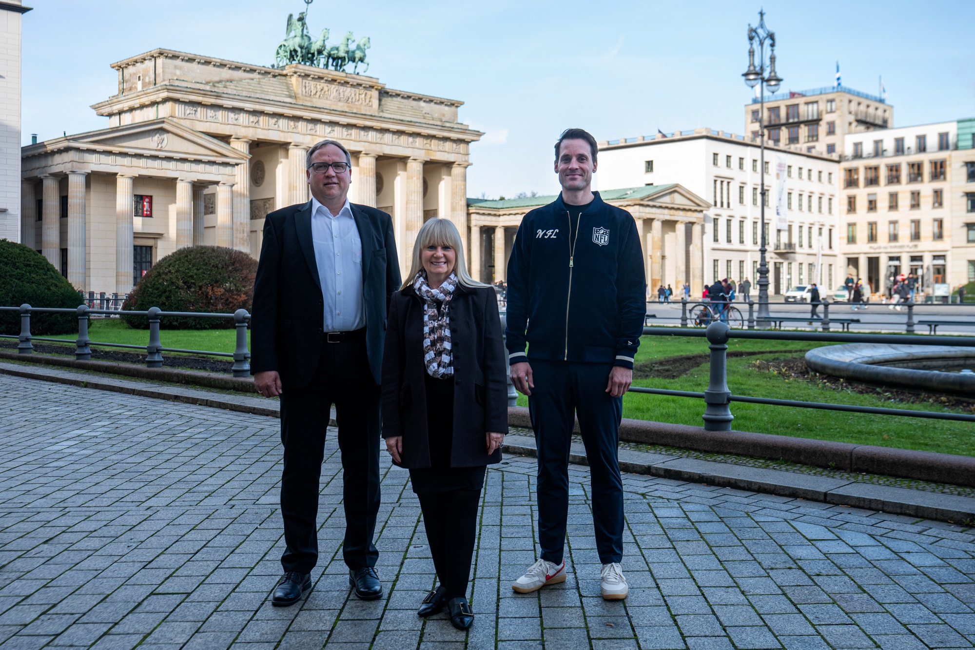 Drei Personen stehen nebeneinander auf einer gepflasterten Fläche in Berlin, mit dem berühmten Brandenburger Tor im Rücken. Sie lächeln und sind sowohl formell als auch leger gekleidet. Es scheint, als würden sie eine der lebhaften Veranstaltungen der Stadt unter einem klaren Himmel genießen. Diese Beschreibung wurde mit der FootballR KI automatisch generiert.