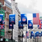 NFL London - Eine Straße in der Stadt ist mit Bannern für die NFL-Spiele in London, britischen und amerikanischen Flaggen und Schildern für NFL-Übertragungen auf BBC und Sky Sports geschmückt. Elegante Gebäude säumen beide Seiten, auf der einen steht ein Baugerüst, das die Begeisterung der britischen Superstars einfängt. Diese Beschreibung wurde mit der FootballR KI automatisch generiert.
