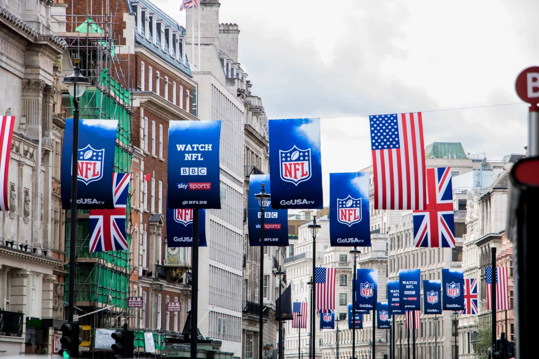 NFL London - Eine Straße in der Stadt ist mit Bannern für die NFL-Spiele in London, britischen und amerikanischen Flaggen und Schildern für NFL-Übertragungen auf BBC und Sky Sports geschmückt. Elegante Gebäude säumen beide Seiten, auf der einen steht ein Baugerüst, das die Begeisterung der britischen Superstars einfängt. Diese Beschreibung wurde mit der FootballR KI automatisch generiert.