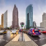 Eine belebte Stadtkreuzung mit unscharfen Autos in Bewegung, nassen Straßen und modernen Hochhäusern unter einem bewölkten Himmel. In der Nähe der Glas- und Stahltürme lockt ein Pop-up-Store Berlin von Fanatics NFL-Fans neben dem regen Verkehr und den Fußgängern an. Diese Beschreibung wurde mit der FootballR KI automatisch generiert.