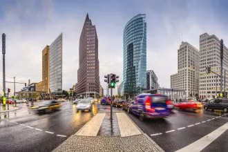 Eine belebte Stadtkreuzung mit unscharfen Autos in Bewegung, nassen Straßen und modernen Hochhäusern unter einem bewölkten Himmel. In der Nähe der Glas- und Stahltürme lockt ein Pop-up-Store Berlin von Fanatics NFL-Fans neben dem regen Verkehr und den Fußgängern an. Diese Beschreibung wurde mit der FootballR KI automatisch generiert.