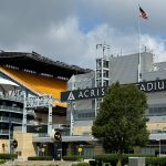 Ein Blick auf das Acrisure Stadion bei bewölktem Himmel, mit dem markanten schwarzen Stadionschild, gelben Sitztribünen, einer amerikanischen Flagge an einem hohen Mast, moderner Architektur, Treppen und einem großen grünen Baum am üppigen Rasen - der Heimat der Pittsburgh Steelers. Diese Beschreibung wurde mit der FootballR KI automatisch generiert.