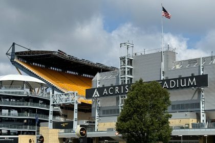 Ein Blick auf das Acrisure Stadion bei bewölktem Himmel, mit dem markanten schwarzen Stadionschild, gelben Sitztribünen, einer amerikanischen Flagge an einem hohen Mast, moderner Architektur, Treppen und einem großen grünen Baum am üppigen Rasen - der Heimat der Pittsburgh Steelers. Diese Beschreibung wurde mit der FootballR KI automatisch generiert.