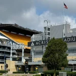Ein Blick auf das Acrisure Stadion bei bewölktem Himmel, mit dem markanten schwarzen Stadionschild, gelben Sitztribünen, einer amerikanischen Flagge an einem hohen Mast, moderner Architektur, Treppen und einem großen grünen Baum am üppigen Rasen - der Heimat der Pittsburgh Steelers. Diese Beschreibung wurde mit der FootballR KI automatisch generiert.