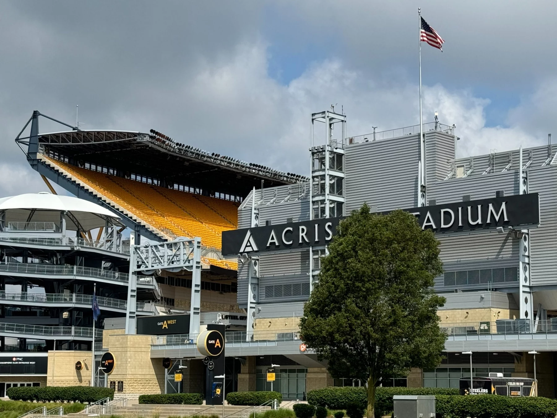 Ein Blick auf das Acrisure Stadion bei bewölktem Himmel, mit dem markanten schwarzen Stadionschild, gelben Sitztribünen, einer amerikanischen Flagge an einem hohen Mast, moderner Architektur, Treppen und einem großen grünen Baum am üppigen Rasen - der Heimat der Pittsburgh Steelers. Diese Beschreibung wurde mit der FootballR KI automatisch generiert.