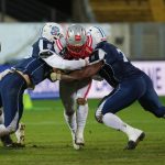 Zwei Football-Spieler in marineblauen Trikots gehen auf einer Wiese gegen einen Spieler in einem weiß-roten Trikot vor, alle tragen Helme und Schützer. Zu sehen sind Spieler des österreichischen Football-Nationalteams, während Headcoach Max Sommer zuschaut. Diese Beschreibung wurde mit der FootballR KI automatisch generiert.