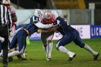 Zwei Football-Spieler in marineblauen Trikots gehen auf einer Wiese gegen einen Spieler in einem weiß-roten Trikot vor, alle tragen Helme und Schützer. Zu sehen sind Spieler des österreichischen Football-Nationalteams, während Headcoach Max Sommer zuschaut. Diese Beschreibung wurde mit der FootballR KI automatisch generiert.