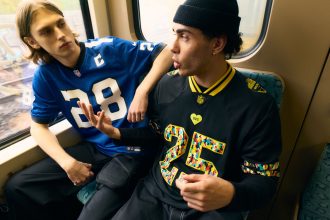Zwei junge Männer sitzen in einem Zug. Der Mann links, in einem blauen NFL-Fan-Shirt mit der Nummer 28, hat lange blonde Haare. Rechts der Mann mit der schwarzen Mütze und dem Berlin Game Trikot mit der Nummer 25, der lebhaft gestikuliert, während er spricht. Diese Beschreibung wurde mit der FootballR KI automatisch generiert.