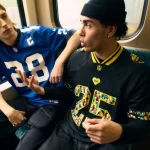 Zwei junge Männer sitzen in einem Zug. Der Mann links, in einem blauen NFL-Fan-Shirt mit der Nummer 28, hat lange blonde Haare. Rechts der Mann mit der schwarzen Mütze und dem Berlin Game Trikot mit der Nummer 25, der lebhaft gestikuliert, während er spricht. Diese Beschreibung wurde mit der FootballR KI automatisch generiert.