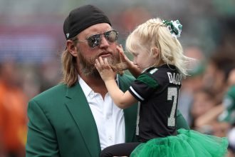 Ex-Jets-Star Nick Mangold, in einer grünen Anzugsjacke, lächelt, als ein junges Mädchen in einem grünen Tutu und einem schwarzen Sporttrikot sein Gesicht berührt. Sie befinden sich im Freien bei einer scheinbaren Sportveranstaltung, umgeben von unscharfen Zuschauern. Diese Beschreibung wurde mit der FootballR KI automatisch generiert.