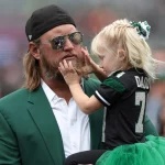 Ex-Jets-Star Nick Mangold, in einer grünen Anzugsjacke, lächelt, als ein junges Mädchen in einem grünen Tutu und einem schwarzen Sporttrikot sein Gesicht berührt. Sie befinden sich im Freien bei einer scheinbaren Sportveranstaltung, umgeben von unscharfen Zuschauern. Diese Beschreibung wurde mit der FootballR KI automatisch generiert.