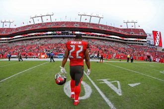 Ein Footballspieler, Doug Martin, trägt ein rotes Trikot der Tampa Bay Buccaneers mit der Nummer 22. Er läuft allein auf dem Spielfeld und hält seinen Helm in der Hand, während die roten Sitze des Stadions und die spärlichen Zuschauer unter dem bedeckten Himmel zu sehen sind. Diese Beschreibung wurde mit der FootballR KI automatisch generiert.