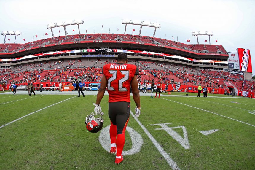 Ein Footballspieler, Doug Martin, trägt ein rotes Trikot der Tampa Bay Buccaneers mit der Nummer 22. Er läuft allein auf dem Spielfeld und hält seinen Helm in der Hand, während die roten Sitze des Stadions und die spärlichen Zuschauer unter dem bedeckten Himmel zu sehen sind. Diese Beschreibung wurde mit der FootballR KI automatisch generiert.
