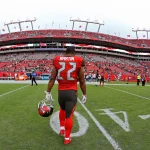 Ein Footballspieler, Doug Martin, trägt ein rotes Trikot der Tampa Bay Buccaneers mit der Nummer 22. Er läuft allein auf dem Spielfeld und hält seinen Helm in der Hand, während die roten Sitze des Stadions und die spärlichen Zuschauer unter dem bedeckten Himmel zu sehen sind. Diese Beschreibung wurde mit der FootballR KI automatisch generiert.