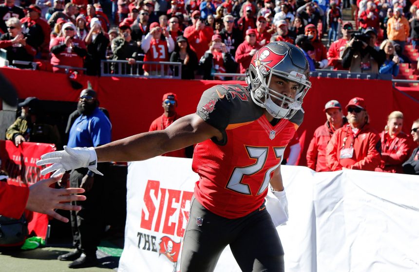 Ein Footballspieler der Tampa Bay Buccaneers, Doug Martin, in einem roten Trikot mit der Nummer 22 streckt seinen Arm aus, um die Fans zu begrüßen. Er befindet sich auf dem Spielfeld mit jubelnden Fans und Sicherheitskräften im Hintergrund, und hinter ihm ist ein großes "Siege the Day"-Banner zu sehen. Diese Beschreibung wurde mit der FootballR KI automatisch generiert.