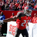 Ein Footballspieler der Tampa Bay Buccaneers, Doug Martin, in einem roten Trikot mit der Nummer 22 streckt seinen Arm aus, um die Fans zu begrüßen. Er befindet sich auf dem Spielfeld mit jubelnden Fans und Sicherheitskräften im Hintergrund, und hinter ihm ist ein großes "Siege the Day"-Banner zu sehen. Diese Beschreibung wurde mit der FootballR KI automatisch generiert.
