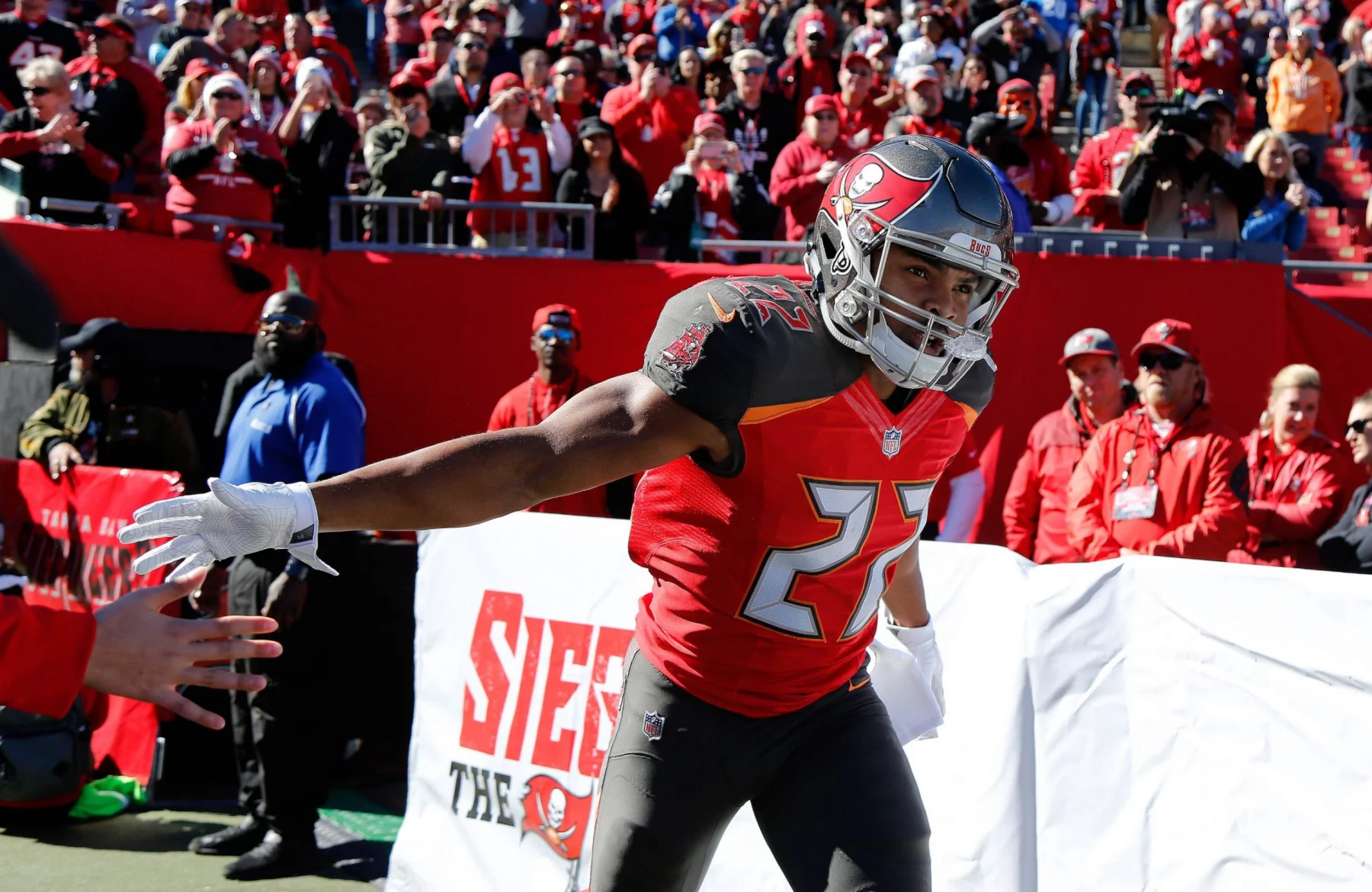Ein Footballspieler der Tampa Bay Buccaneers, Doug Martin, in einem roten Trikot mit der Nummer 22 streckt seinen Arm aus, um die Fans zu begrüßen. Er befindet sich auf dem Spielfeld mit jubelnden Fans und Sicherheitskräften im Hintergrund, und hinter ihm ist ein großes "Siege the Day"-Banner zu sehen. Diese Beschreibung wurde mit der FootballR KI automatisch generiert.