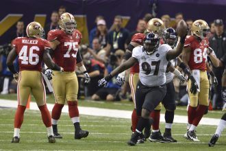 Der Spieler der Baltimore Ravens, Arthur Jones (Nummer 97), jubelt mit einem Football in der Hand während eines NFL-Super-Bowl-Spiels gegen die San Francisco 49ers, während Spieler in roten Trikots in der Nähe des Spielfelds stehen und Zuschauer im Hintergrund zu sehen sind. Diese Beschreibung wurde mit der FootballR KI automatisch generiert.