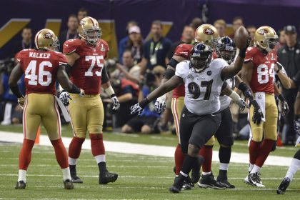 Der Spieler der Baltimore Ravens, Arthur Jones (Nummer 97), jubelt mit einem Football in der Hand während eines NFL-Super-Bowl-Spiels gegen die San Francisco 49ers, während Spieler in roten Trikots in der Nähe des Spielfelds stehen und Zuschauer im Hintergrund zu sehen sind. Diese Beschreibung wurde mit der FootballR KI automatisch generiert.