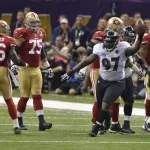 Der Spieler der Baltimore Ravens, Arthur Jones (Nummer 97), jubelt mit einem Football in der Hand während eines NFL-Super-Bowl-Spiels gegen die San Francisco 49ers, während Spieler in roten Trikots in der Nähe des Spielfelds stehen und Zuschauer im Hintergrund zu sehen sind. Diese Beschreibung wurde mit der FootballR KI automatisch generiert.