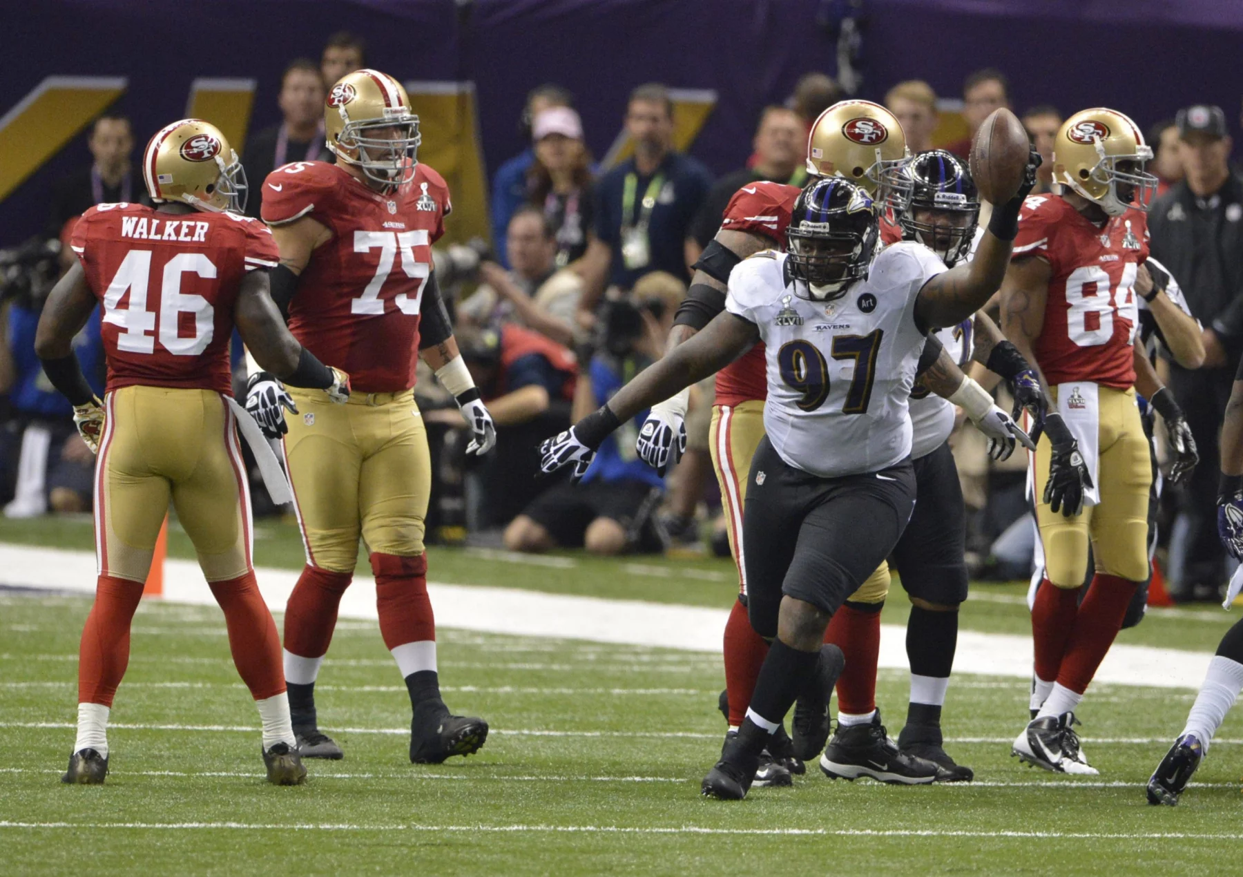 Der Spieler der Baltimore Ravens, Arthur Jones (Nummer 97), jubelt mit einem Football in der Hand während eines NFL-Super-Bowl-Spiels gegen die San Francisco 49ers, während Spieler in roten Trikots in der Nähe des Spielfelds stehen und Zuschauer im Hintergrund zu sehen sind. Diese Beschreibung wurde mit der FootballR KI automatisch generiert.
