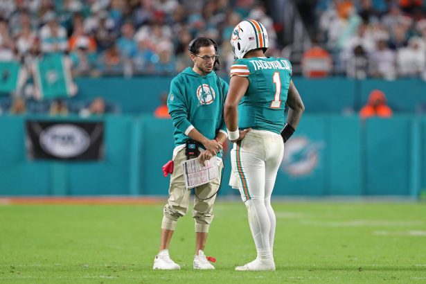 Miami Dolphins-Coach Mike McDaniel im blauen Kapuzenpulli spricht mit Quarterback Tua Tagovailoa (#1) auf dem Spielfeld. Beide wirken konzentriert, während McDaniel ein Spielblatt in der Hand hält. Das Stadion und die unscharfe, in Grüntöne getauchte Menge bilden die Kulisse. Diese Beschreibung wurde mit der FootballR KI automatisch generiert.