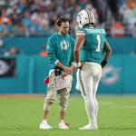 Miami Dolphins-Coach Mike McDaniel im blauen Kapuzenpulli spricht mit Quarterback Tua Tagovailoa (#1) auf dem Spielfeld. Beide wirken konzentriert, während McDaniel ein Spielblatt in der Hand hält. Das Stadion und die unscharfe, in Grüntöne getauchte Menge bilden die Kulisse. Diese Beschreibung wurde mit der FootballR KI automatisch generiert.