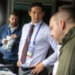 Vier Männer stehen in einer Sendekabine. Ex-Jets-Quarterback Mark Sanchez, in weißem Hemd und Krawatte mit einem Schlüsselband, blickt nach links. Zwei Männer in legeren Jacken stehen in der Nähe - einer mit einem Notizblock -, während ein anderer Mann in Blau dahinter steht und beobachtet. Diese Beschreibung wurde mit der FootballR KI automatisch generiert.