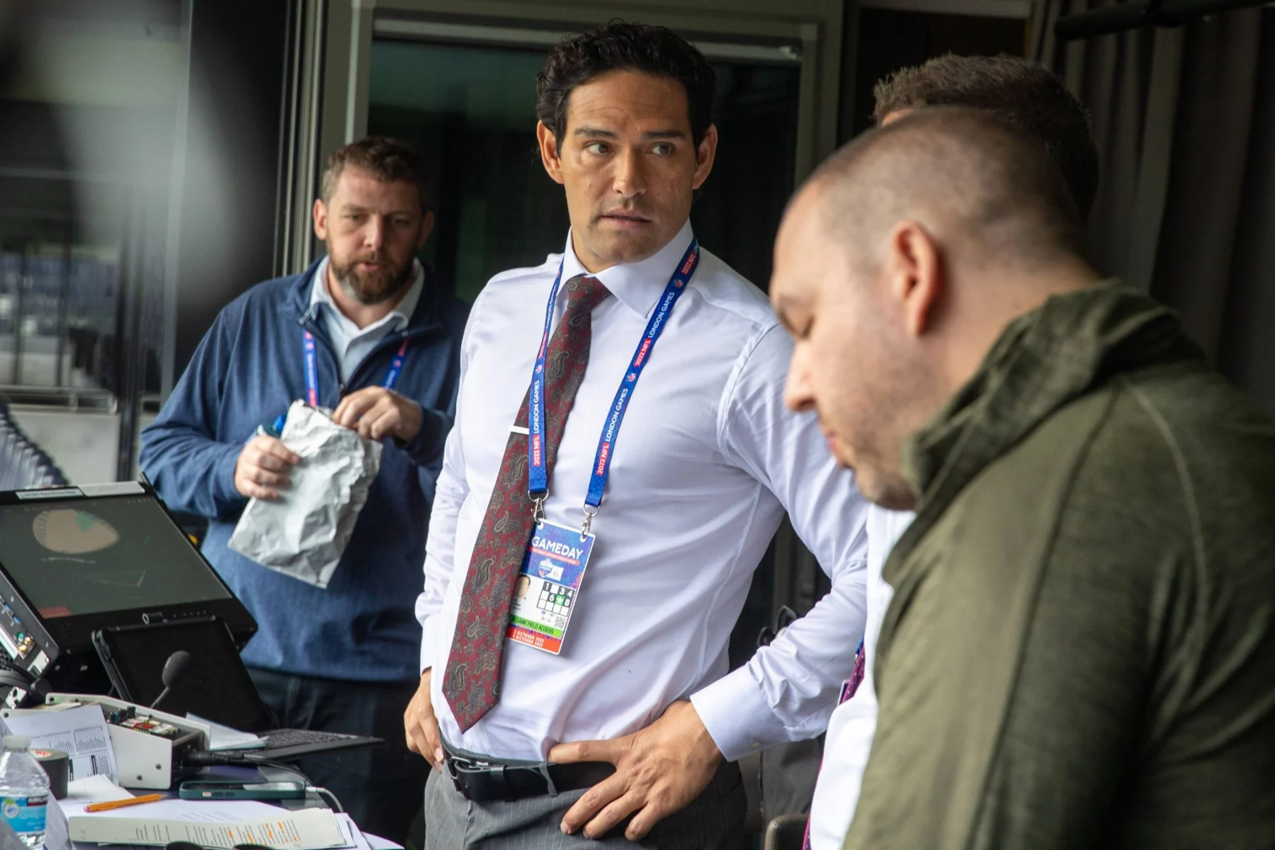 Vier Männer stehen in einer Sendekabine. Ex-Jets-Quarterback Mark Sanchez, in weißem Hemd und Krawatte mit einem Schlüsselband, blickt nach links. Zwei Männer in legeren Jacken stehen in der Nähe - einer mit einem Notizblock -, während ein anderer Mann in Blau dahinter steht und beobachtet. Diese Beschreibung wurde mit der FootballR KI automatisch generiert.