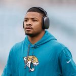 Tyson Campbell - Ein Footballspieler in einem blaugrünen Jaguars-Kapuzenpulli mit dem Jaguar-Logo der Mannschaft steht mit großen schwarzen Kopfhörern im Freien. Der Cornerback blickt mit ernster Miene nach vorne, während der verschwommene Hintergrund die Aufmerksamkeit auf ihn lenkt. Diese Beschreibung wurde mit der FootballR KI automatisch generiert.