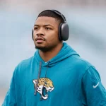 Tyson Campbell - Ein Footballspieler in einem blaugrünen Jaguars-Kapuzenpulli mit dem Jaguar-Logo der Mannschaft steht mit großen schwarzen Kopfhörern im Freien. Der Cornerback blickt mit ernster Miene nach vorne, während der verschwommene Hintergrund die Aufmerksamkeit auf ihn lenkt. Diese Beschreibung wurde mit der FootballR KI automatisch generiert.