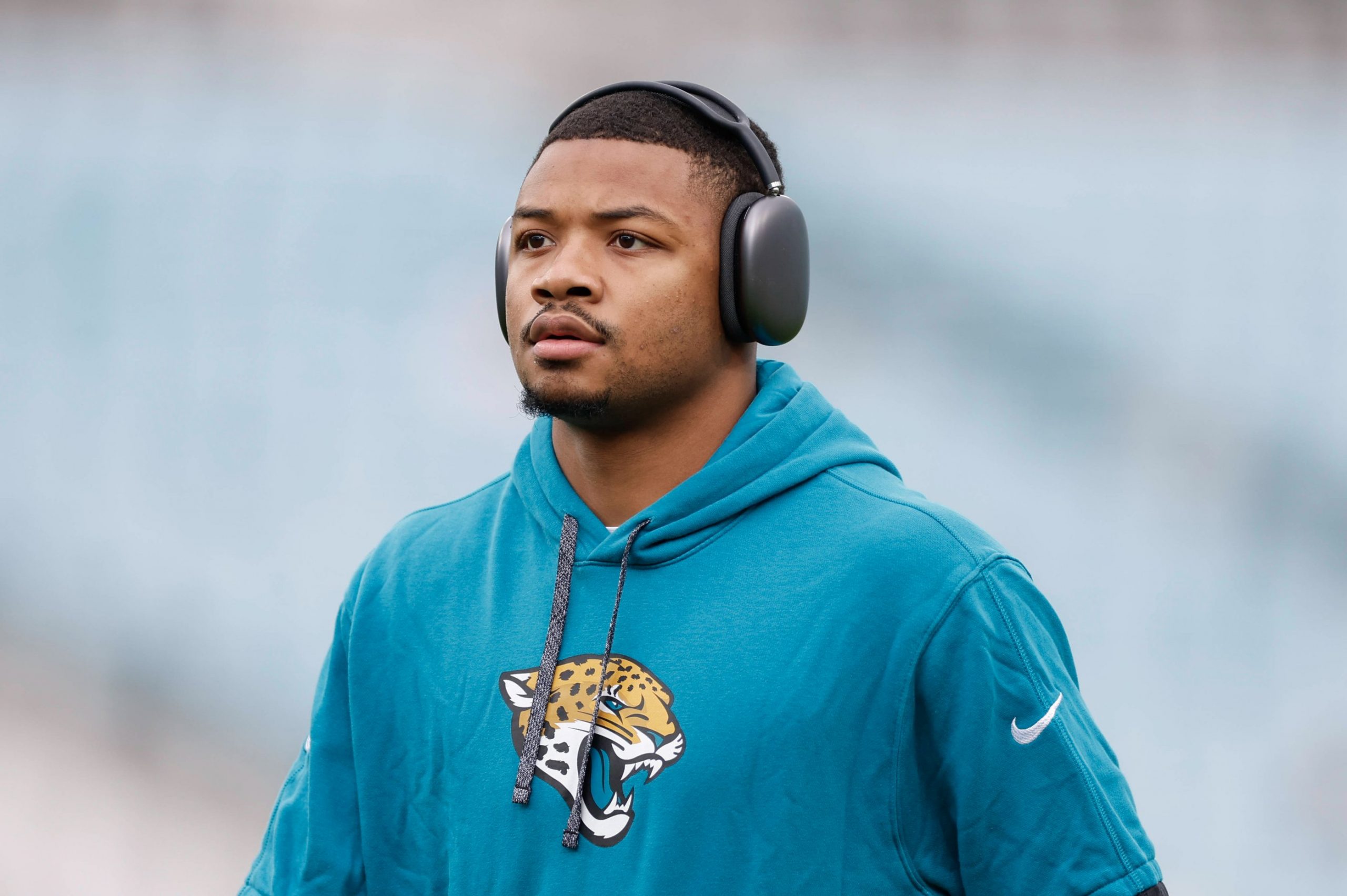 Tyson Campbell - Ein Footballspieler in einem blaugrünen Jaguars-Kapuzenpulli mit dem Jaguar-Logo der Mannschaft steht mit großen schwarzen Kopfhörern im Freien. Der Cornerback blickt mit ernster Miene nach vorne, während der verschwommene Hintergrund die Aufmerksamkeit auf ihn lenkt. Diese Beschreibung wurde mit der FootballR KI automatisch generiert.