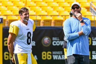 Aaron Rodgers und Ben Roethlisberger. Ein lächelnder Football-Spieler in einem Steelers-Trikot mit der Nummer 8 steht auf dem Spielfeld und hält seinen Helm. Neben ihm steht ein bärtiger Mann mit Sonnenbrille und Mütze. Beide stehen entspannt in der Nähe der gelben Stadionsitze und denken über das Erreichen eines historischen Meilensteins nach. Diese Beschreibung wurde mit der FootballR KI automatisch generiert.