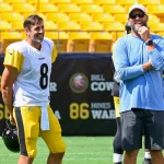 Aaron Rodgers und Ben Roethlisberger. Ein lächelnder Football-Spieler in einem Steelers-Trikot mit der Nummer 8 steht auf dem Spielfeld und hält seinen Helm. Neben ihm steht ein bärtiger Mann mit Sonnenbrille und Mütze. Beide stehen entspannt in der Nähe der gelben Stadionsitze und denken über das Erreichen eines historischen Meilensteins nach. Diese Beschreibung wurde mit der FootballR KI automatisch generiert.