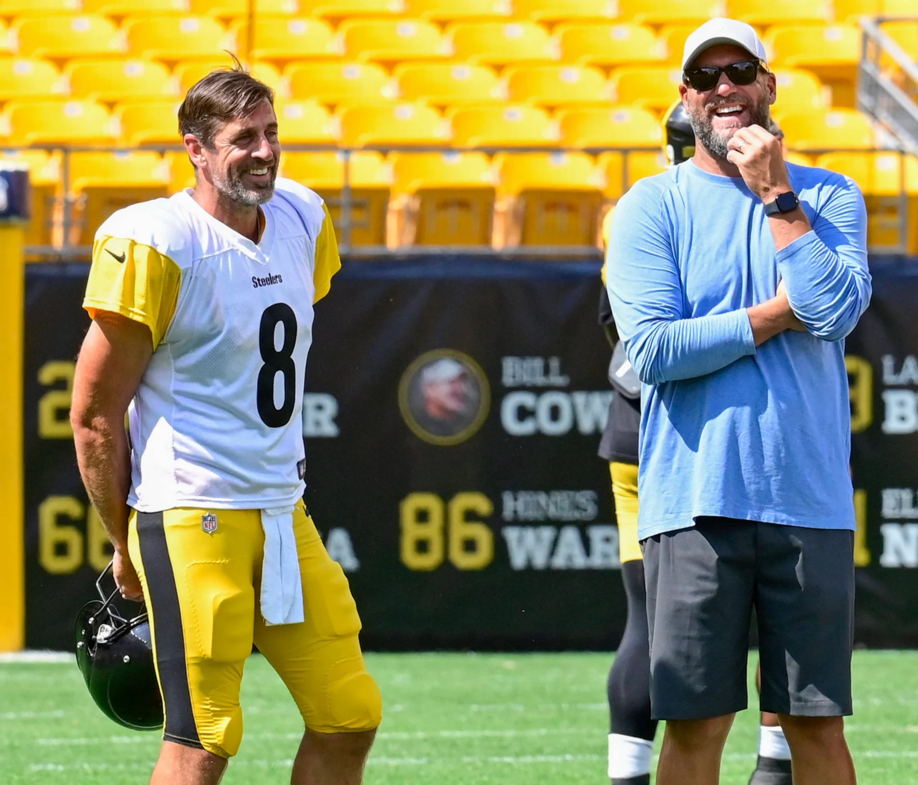 Aaron Rodgers und Ben Roethlisberger. Ein lächelnder Football-Spieler in einem Steelers-Trikot mit der Nummer 8 steht auf dem Spielfeld und hält seinen Helm. Neben ihm steht ein bärtiger Mann mit Sonnenbrille und Mütze. Beide stehen entspannt in der Nähe der gelben Stadionsitze und denken über das Erreichen eines historischen Meilensteins nach. Diese Beschreibung wurde mit der FootballR KI automatisch generiert.