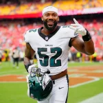 Ein lächelnder NFL-Spieler der Philadelphia Eagles, der die Trikotnummer 52 trägt, hält seinen Helm und zeigt auf dem Spielfeld ein Friedenszeichen, das an den Stil von Za'Darius Smiths Karriere erinnert. Hinter ihm sind verschwommene Stadionsitze zu sehen, die mit Fans in Rot gefüllt sind. Diese Beschreibung wurde mit der FootballR KI automatisch generiert.