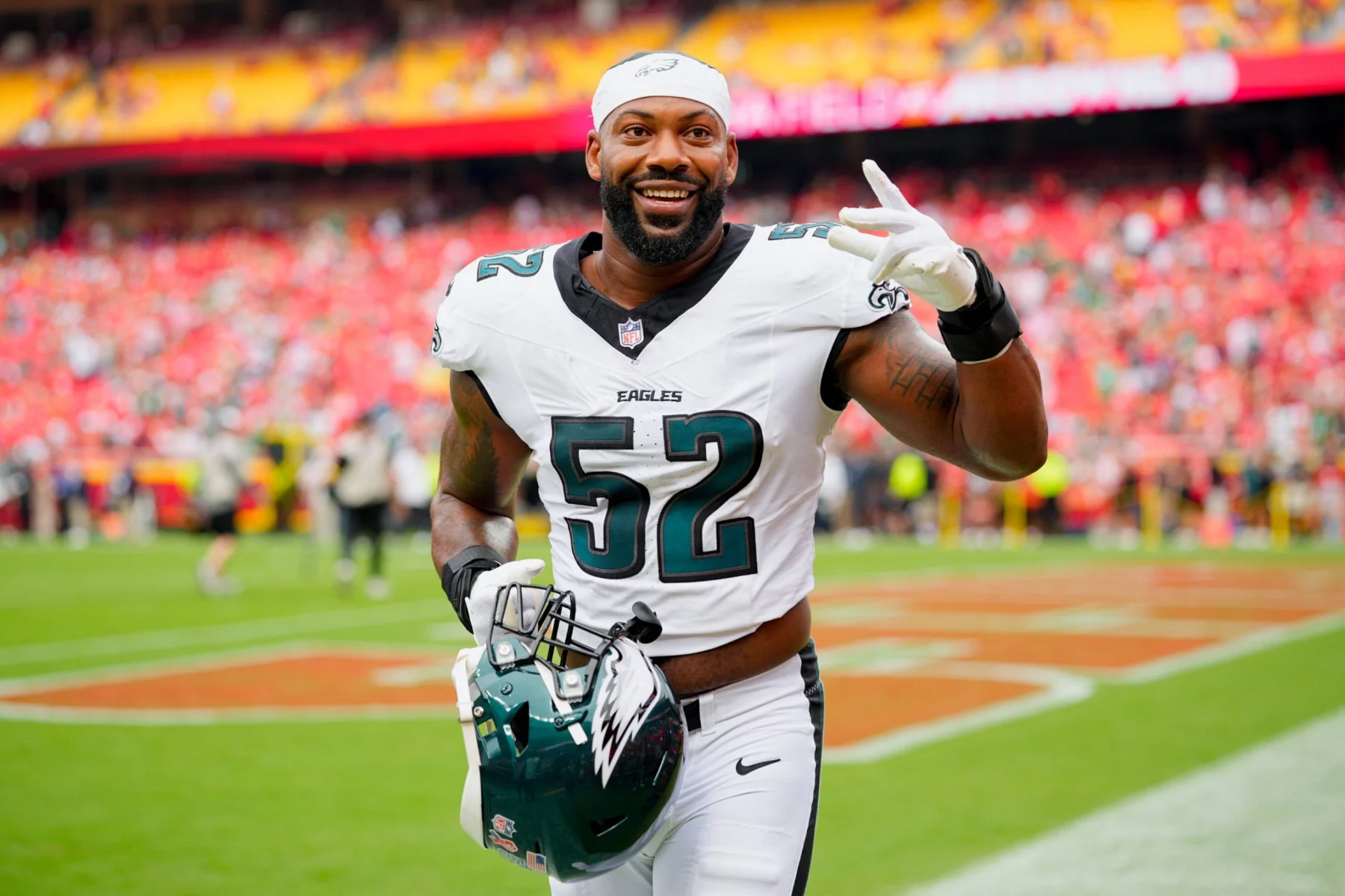 Ein lächelnder NFL-Spieler der Philadelphia Eagles, der die Trikotnummer 52 trägt, hält seinen Helm und zeigt auf dem Spielfeld ein Friedenszeichen, das an den Stil von Za'Darius Smiths Karriere erinnert. Hinter ihm sind verschwommene Stadionsitze zu sehen, die mit Fans in Rot gefüllt sind. Diese Beschreibung wurde mit der FootballR KI automatisch generiert.