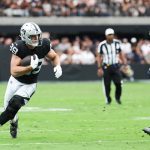 Ein Football-Spieler in einem schwarz-silbernen Raiders-Trikot läuft mit dem Ball und schaut nach vorne, während er von einem Bears-Verteidiger verfolgt wird. Die Aktion ist intensiv, ähnlich wie der herausragende Tight End Brock Bowers, der auf einem leuchtend grünen Feld vor jubelnden Fans spielt. Diese Beschreibung wurde mit der FootballR KI automatisch generiert.