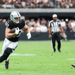 Ein Football-Spieler in einem schwarz-silbernen Raiders-Trikot läuft mit dem Ball und schaut nach vorne, während er von einem Bears-Verteidiger verfolgt wird. Die Aktion ist intensiv, ähnlich wie der herausragende Tight End Brock Bowers, der auf einem leuchtend grünen Feld vor jubelnden Fans spielt. Diese Beschreibung wurde mit der FootballR KI automatisch generiert.