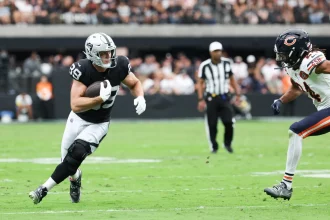 Ein Football-Spieler in einem schwarz-silbernen Raiders-Trikot läuft mit dem Ball und schaut nach vorne, während er von einem Bears-Verteidiger verfolgt wird. Die Aktion ist intensiv, ähnlich wie der herausragende Tight End Brock Bowers, der auf einem leuchtend grünen Feld vor jubelnden Fans spielt. Diese Beschreibung wurde mit der FootballR KI automatisch generiert.