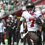 Bucky Irving - Ein Footballspieler der Tampa Bay Buccaneers, der die Trikotnummer 7 und schwarze Handschuhe trägt, läuft während eines Spiels gegen Seattle mit dem Ball. Unscharfe Gegner und Mannschaftskameraden füllen das Spielfeld, und auf den Sitzen im Stadion ist eine jubelnde Menge zu sehen. Diese Beschreibung wurde mit der FootballR KI automatisch generiert.